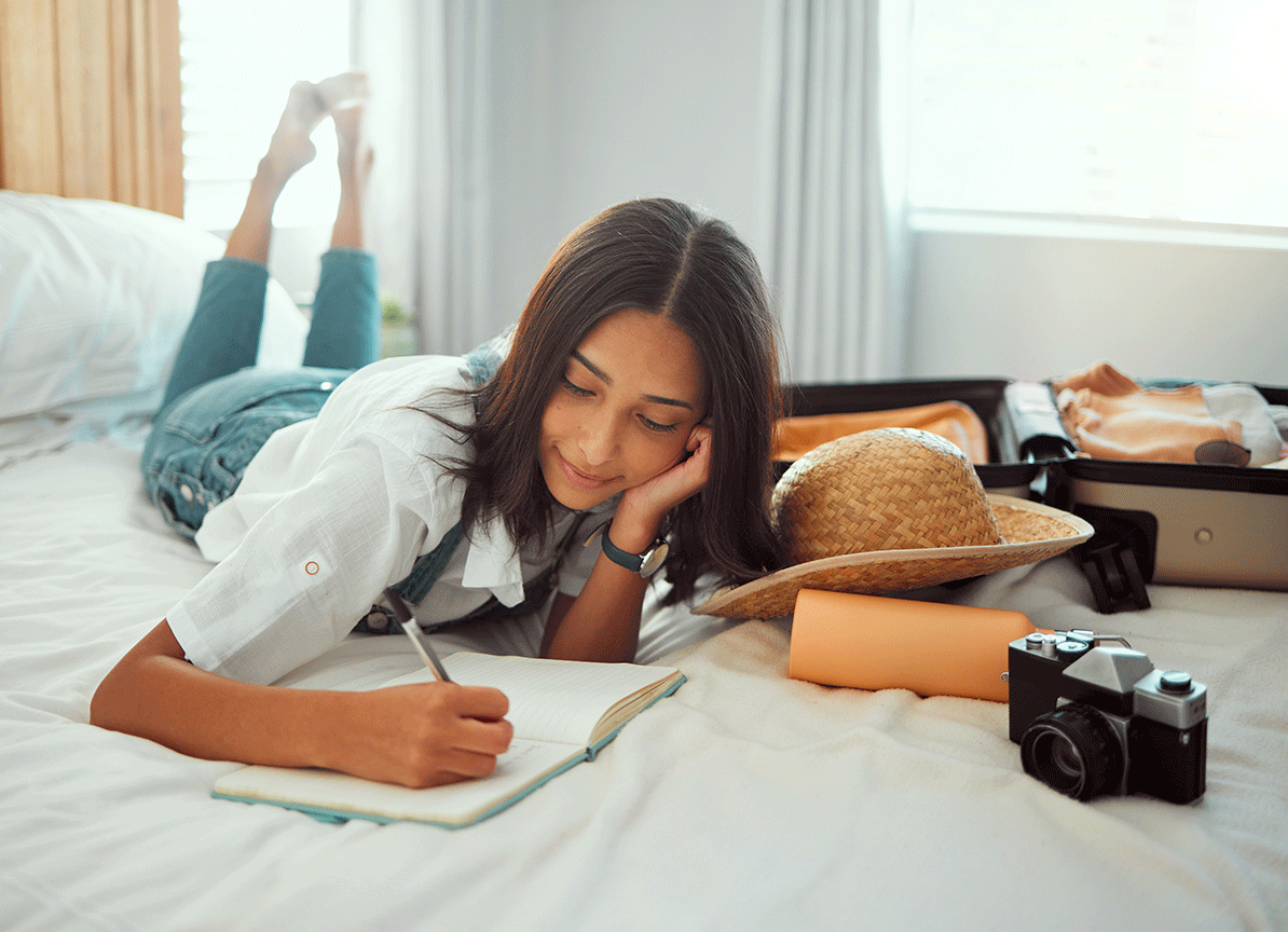 A teenage girl lying on her stomach on a bed writing in a notebook