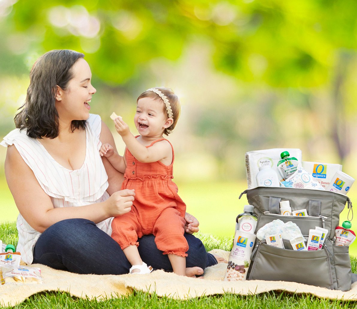 Mom and baby sitting on the grass with a diaper bag filled with Own Brands baby products